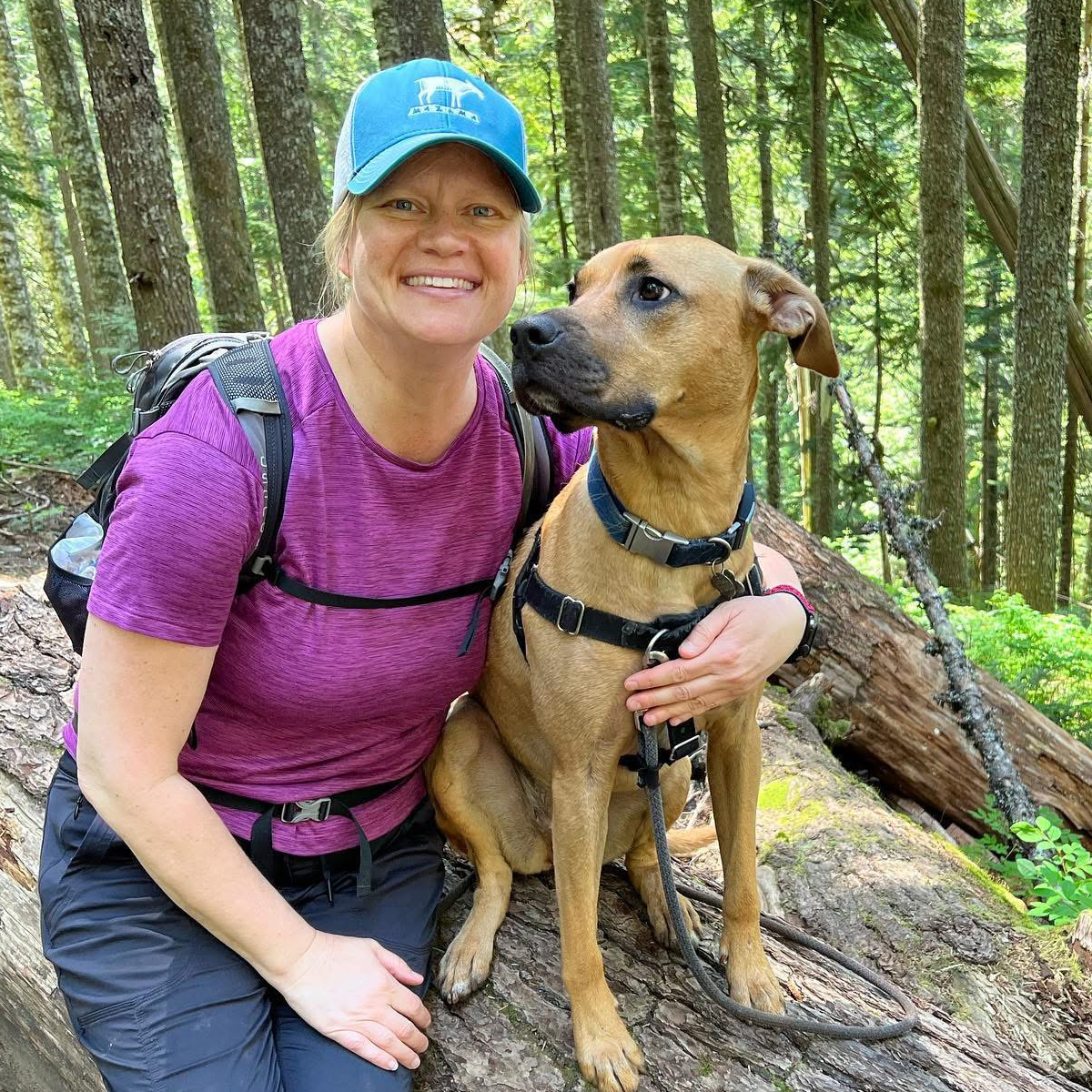 Christa and Rikka smiling for a photo while sitting on a tree log during a hiking adventure.