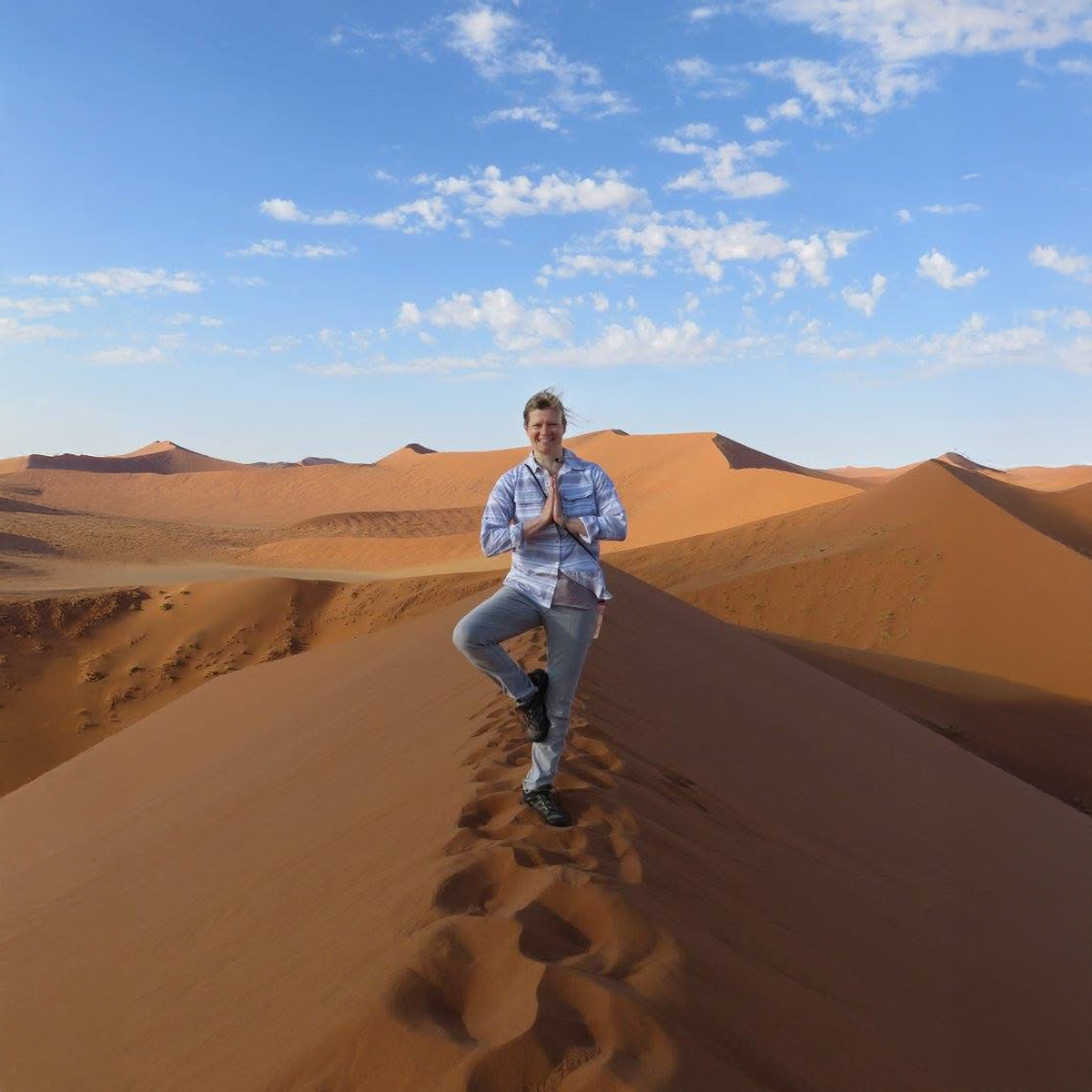 Christa smiling and facing the camera while doing tree pose on the sienna-colored sand dunes of Namibia, under clear blue skies.