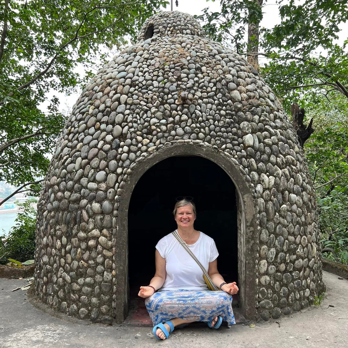 Christa sitting in lotus just inside a rounded, igloo-like structure covered in small stones and pebbles, surrounded by trees at the Beatles Ashram.