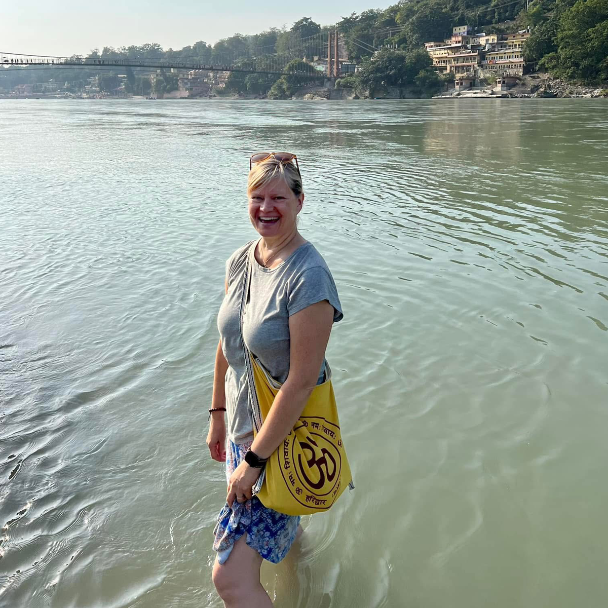 Christa standing in the Ganga River, wearing a floral skirt and carrying a yellow bag, with a bridge and buildings visible in the distance.