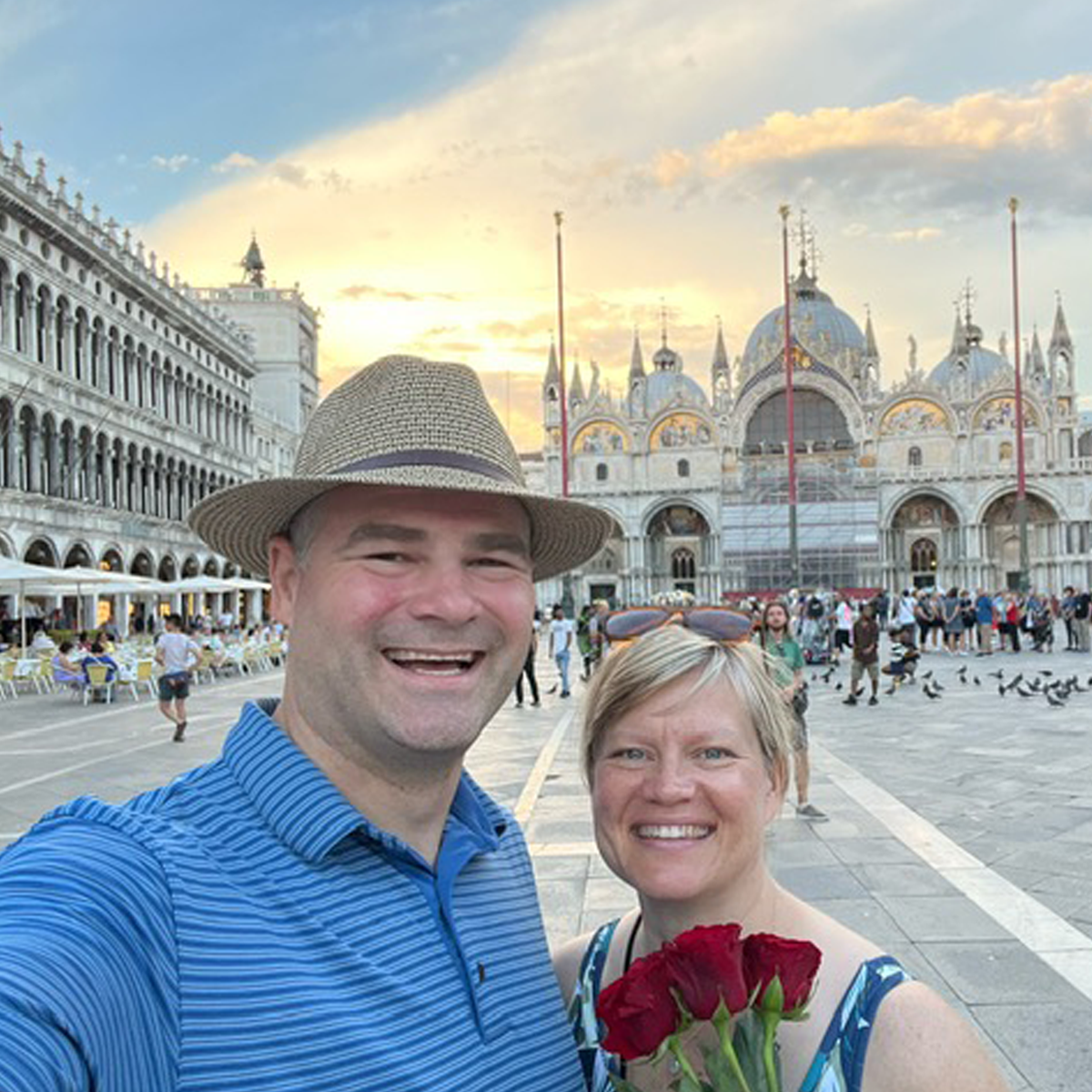 Christa and Mike posing for a selfie in front of St. Mark's Basilica in Venice, with Mike who is wearing a hat and taking the photo and Christa holding red roses; people and pigeons fill the background.